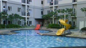 A swimming pool with a slide and a small red slide, surrounded by a building with balconies and greenery. - Cozy Stay 1Br At Maple Park Apartment Near Sunter (Jakarta)