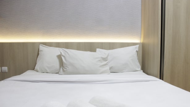 Cama bem arrumada com lençóis e almofadas brancas, cabeceira de madeira e luminária fixada na parede.