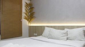 Cama bem arrumada com lençóis e almofadas brancas, luminária fixada na parede e painel de madeira à esquerda.