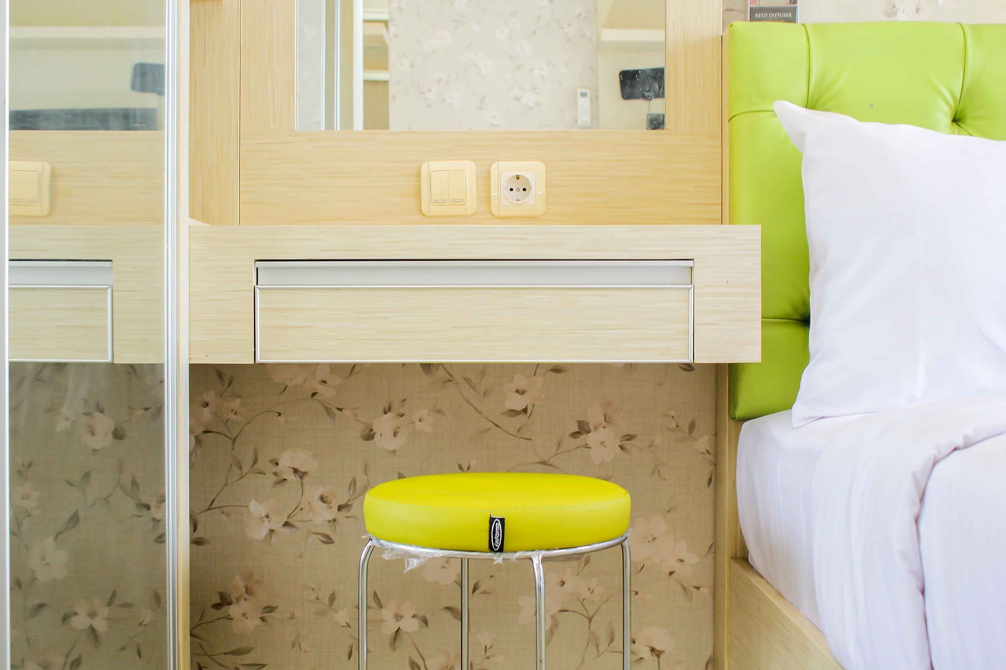 A hotel room with a bed, a green upholstered headboard, a yellow stool, a wooden vanity with a mirror, and a floral wallpaper.