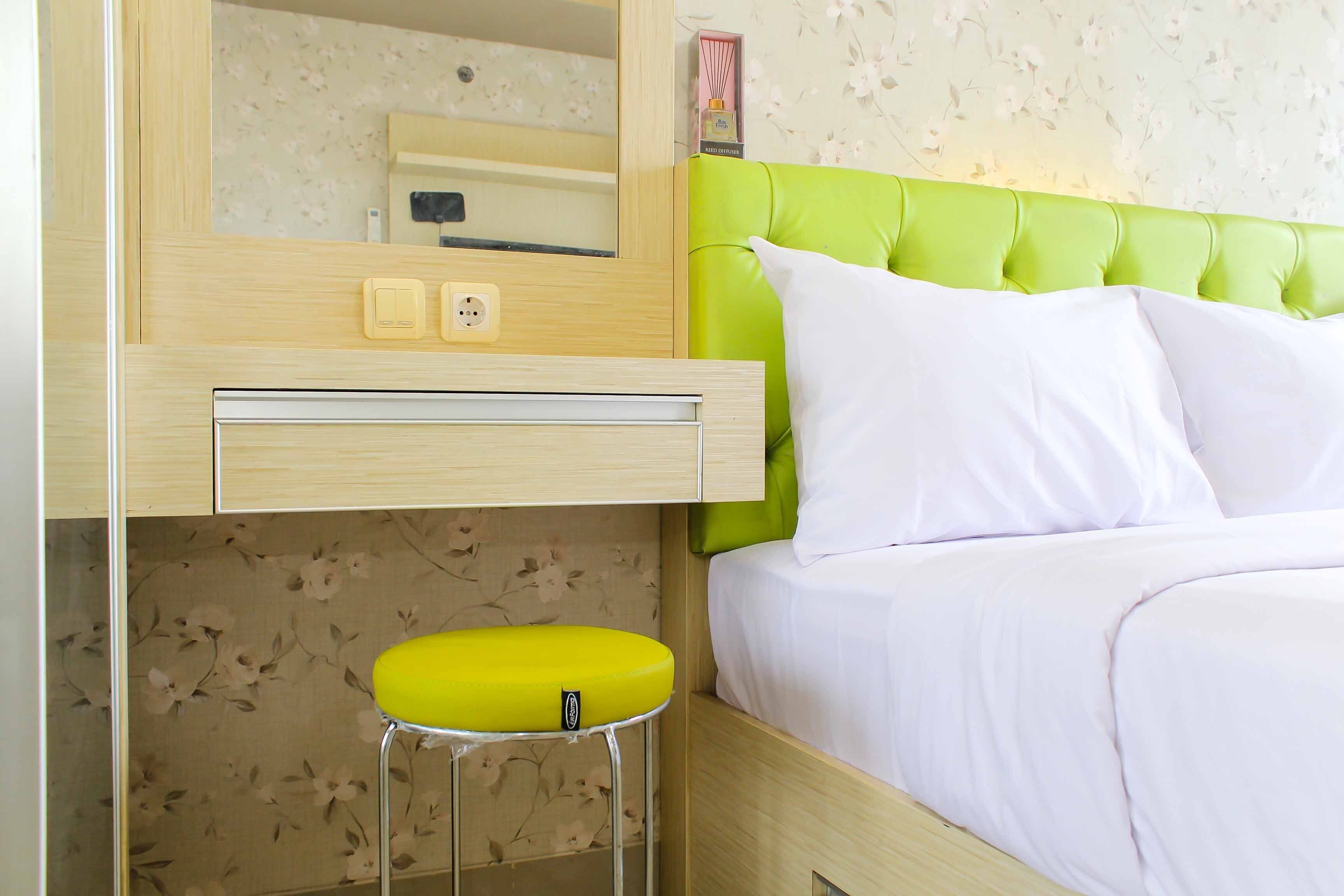 Un dormitorio con cabecera tapizada de verde, una cama blanca, un taburete amarillo y un aparador de madera con espejo.