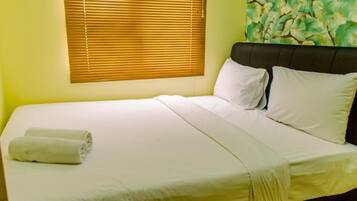 A neatly made bed with white linens and pillows, a rolled towel, and a window with blinds.