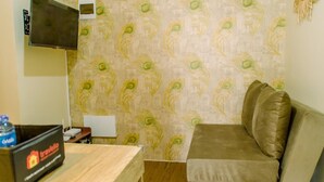 A room with a peacock feather patterned wallpaper, a brown sofa, a television mounted on the wall, and a small table with a water bottle and a box labeled "Travelio".