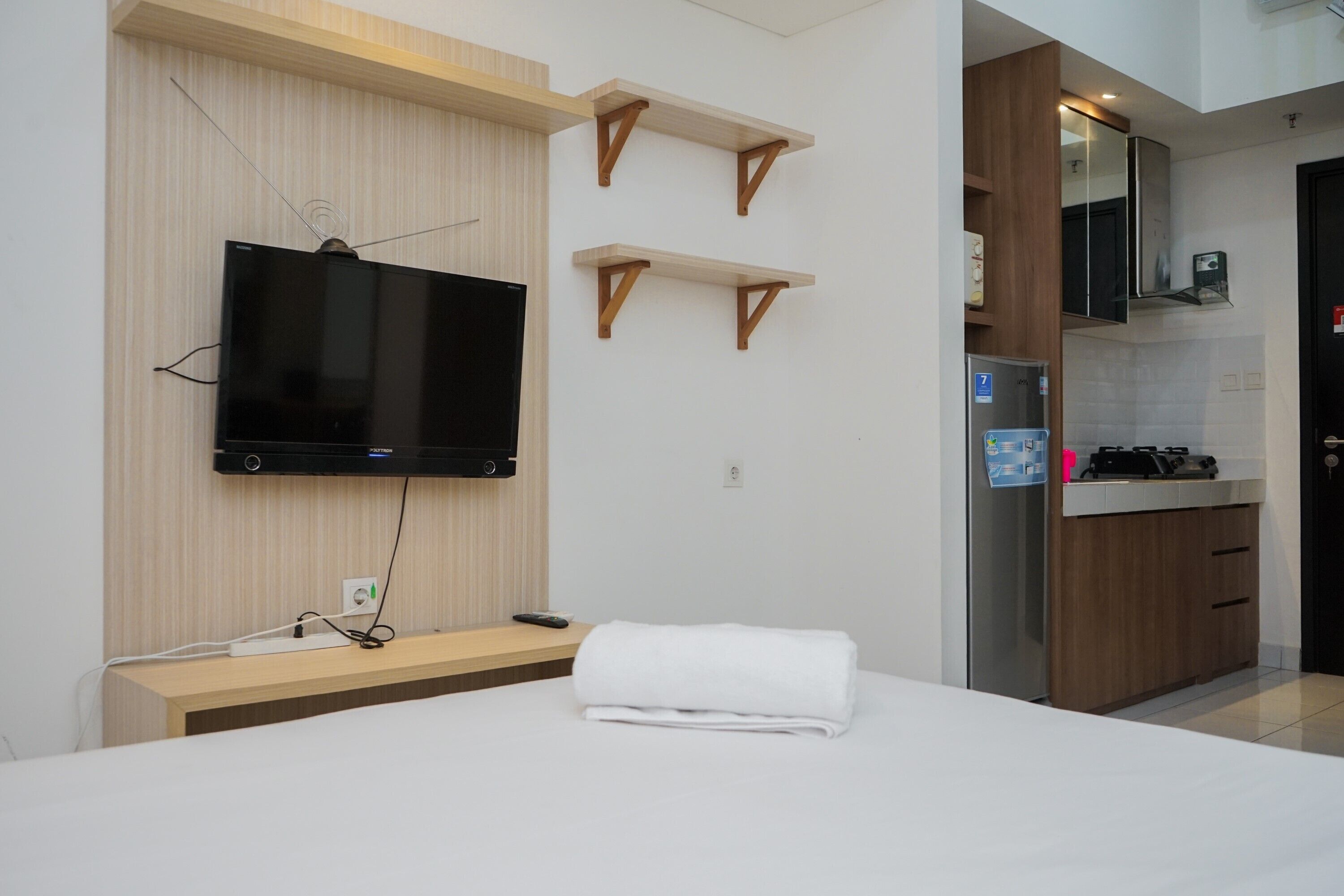 Ett modernt hotellrum med en platt-TV som är monterad på väggen, hyllor i trä, ett köksutrymme med kylskåp och en snyggt bäddad säng med en vit handduk.