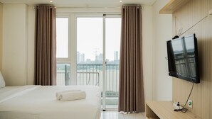 A hotel room with a bed, a television mounted on the wall, and a view of a cityscape through the window. - Minimalist And Warm Studio Casa De Parco Apartment (Pagedangan)