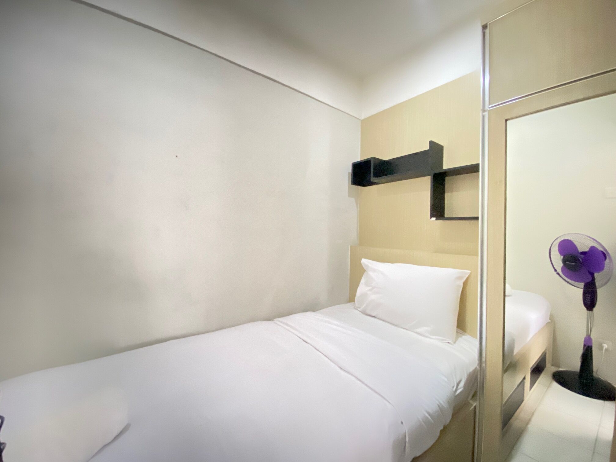 A single bed with white bedding in a room with a beige wall and a small shelf above it.