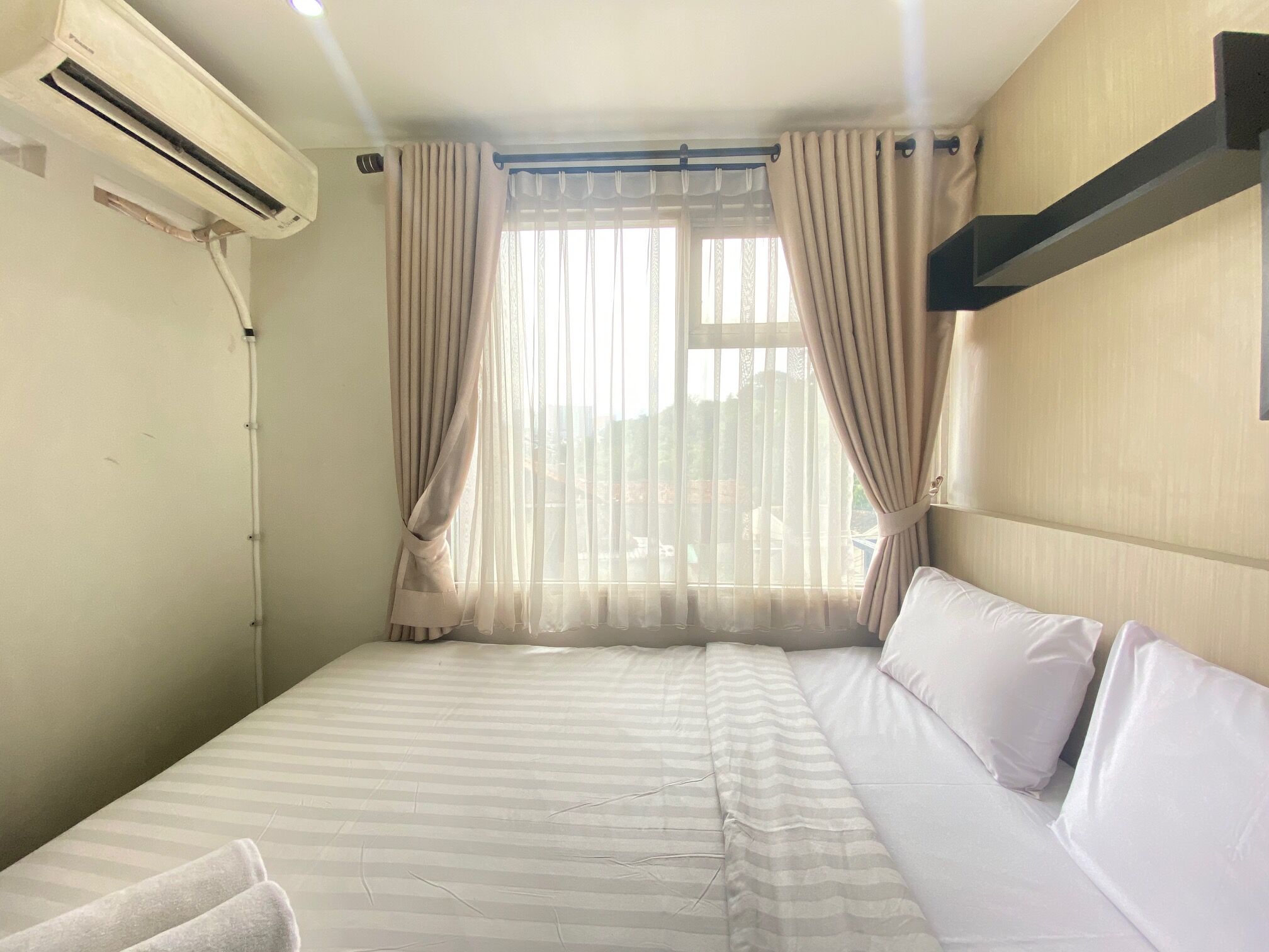 A hotel room with a bed, curtains, an air conditioning unit, and a view of the outside.