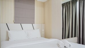 A neatly made bed with white linens and pillows, a headboard with a light wood finish, and a window with dark curtains.