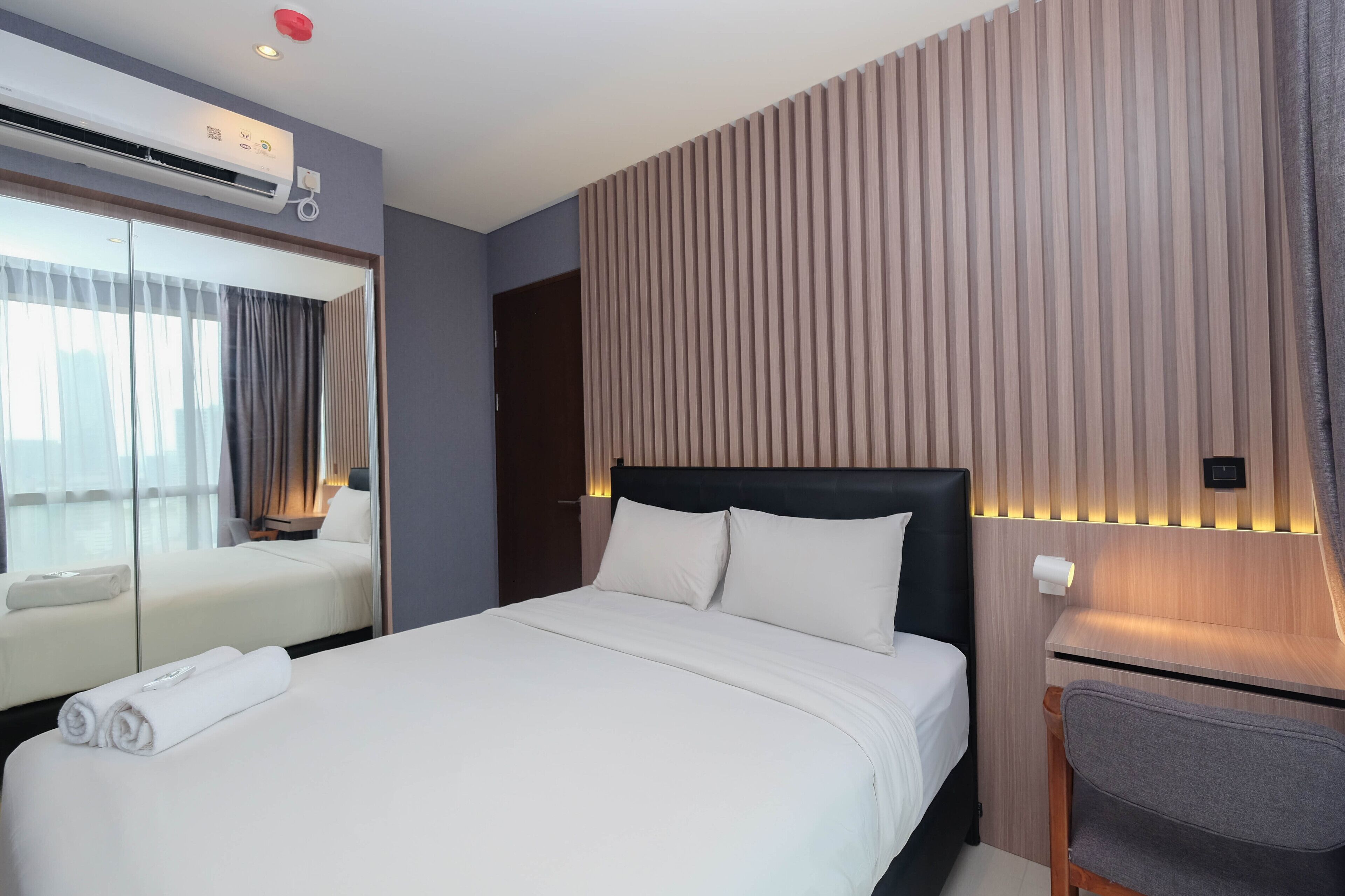 A hotel room with a bed, a desk, a chair, a mirror, and a wall-mounted air conditioner.