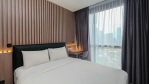A modern hotel room with a large bed, a desk, and a view of the city through sheer curtains.