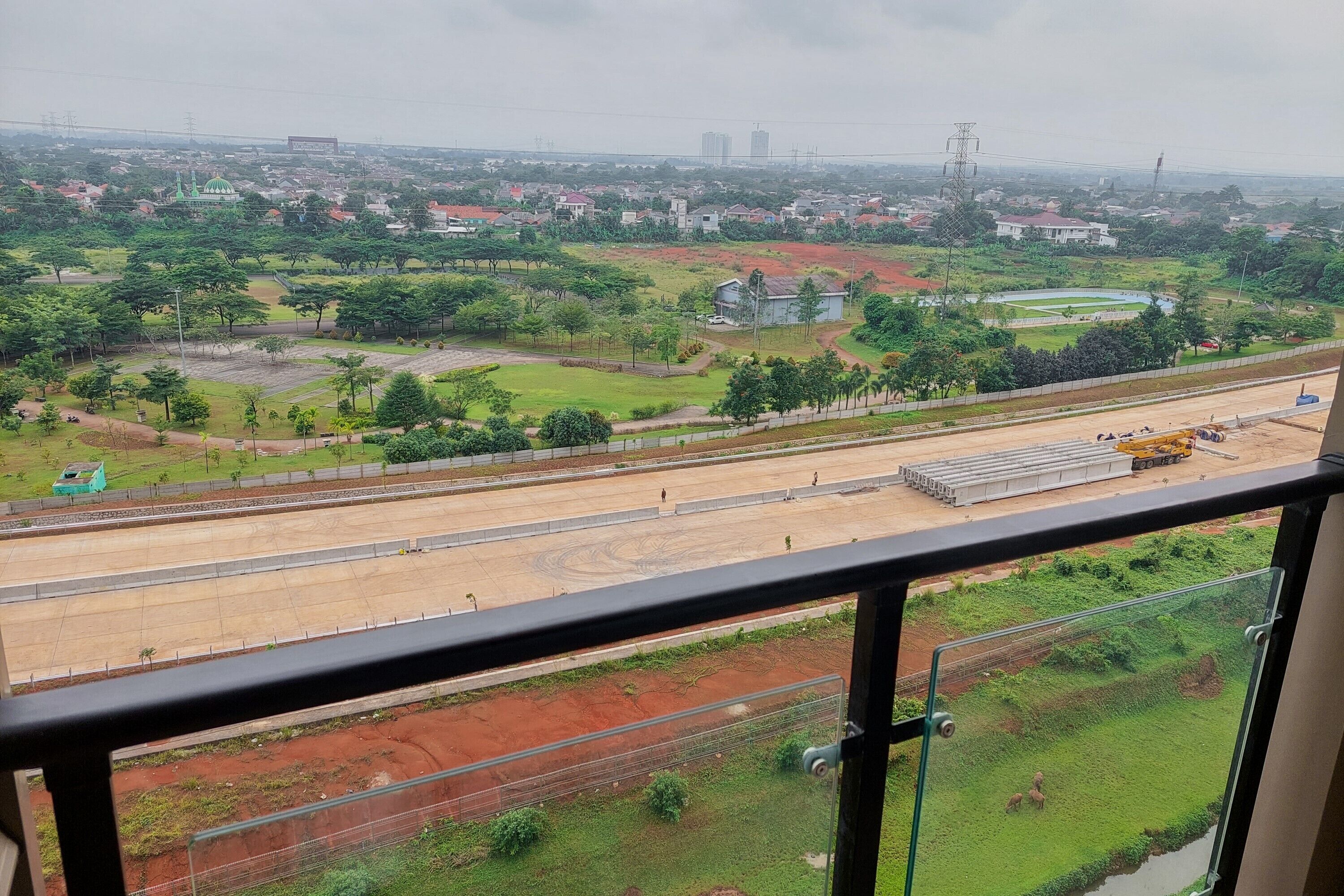 Vista desde un balcón que domina un paisaje urbano con un espacio abierto, áreas verdes y una carretera con vehículos de construcción.