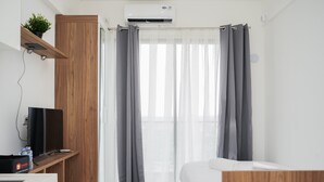 A modern hotel room with a wooden wardrobe, a flat-screen TV, a bedside table, and a large window with grey curtains. - Nice And Elegant Studio Apartment At Sky House Bsd (Pagedangan)