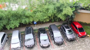 A parking lot with several cars parked in a row, a blue parking sign, and a dense green tree line in the background. - Cozy Stay Studio Apartment (No Kitchen) At Metropark Condominium Jababeka (Cikarang)