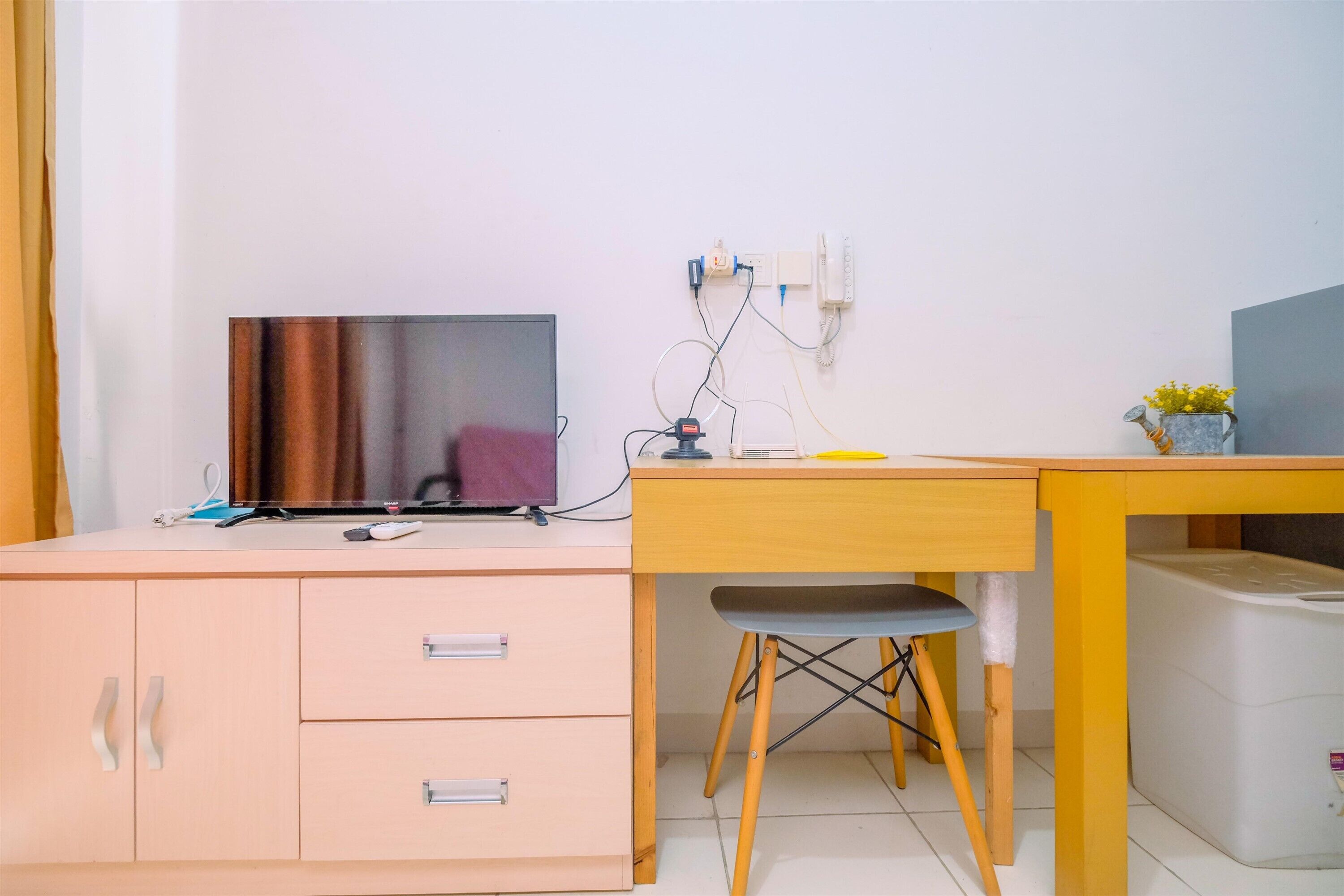 A small, neatly organized room with a desk, a chair, a television, and a small table with a vase of flowers.