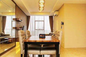 A modern hotel room with a wooden dining table, leather chairs, a chandelier, a sofa, and a large window with curtains.