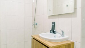 A bathroom with white tiles, a wooden vanity, a sink, a mirror, and a shower head. - Comfy Studio At Amazana Serpong Apartment (South Tangerang)