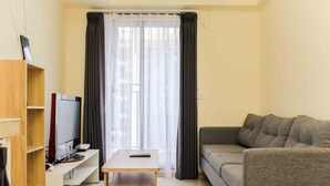 A compact living room with a flat-screen TV on a wooden stand, a grey sofa, and a small table. - Comfort And Serene 2Br At Meikarta Apartment (Cikarang)
