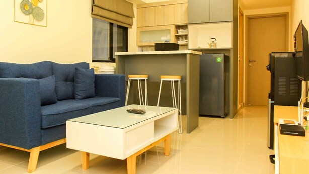 A compact living space with a sofa, a glass-top coffee table, a kitchen area with a refrigerator, and a television. - Comfortable And Spacious 2Br At Meikarta Apartment (Cikarang)