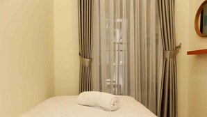 A neatly made bed with a rolled white towel, a wooden shelf with a mirror, and sheer curtains with a window in the background. - Comfortable And Spacious 2Br At Meikarta Apartment (Cikarang)