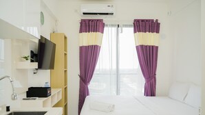 A hotel room with a bed, a TV, a sink, and a window with purple curtains. - Pleasurable Studio At Sky House Bsd Apartment (Pagedangan)
