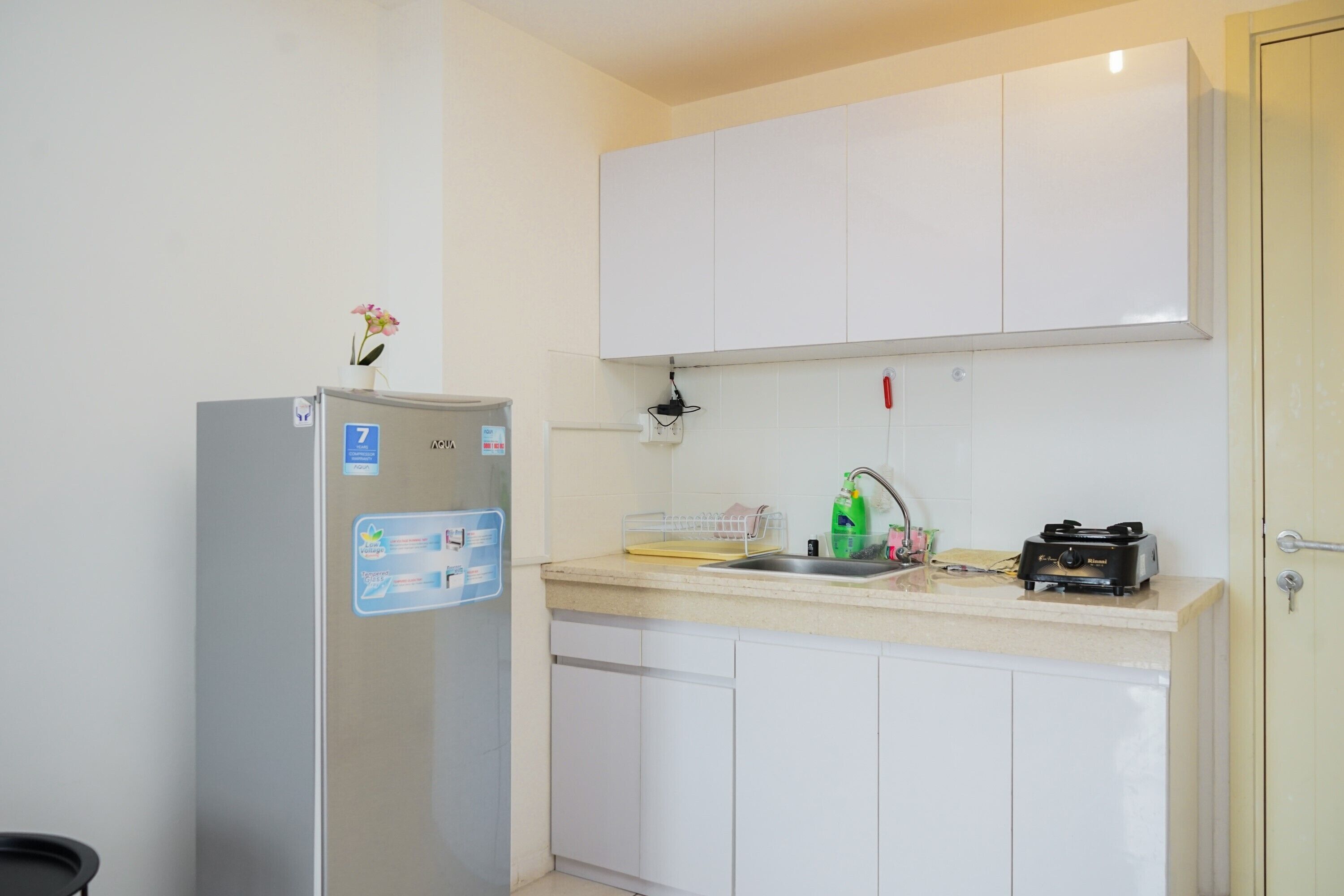 A compact kitchen with white cabinets, a stainless steel refrigerator, a sink with a dish rack, and a small countertop with a kettle and a bottle of dish soap.