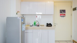 A compact kitchen with white cabinets, a sink, and a refrigerator. There is a fire safety sign on the door and a small plant on the counter. - Restful And Cozy 2Br Apartment At M-Town Residence (Gading Serpong)
