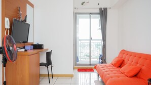 A modern living room with a wooden desk, a computer, a fan, a red rug, and a bright orange sofa. - Restful And Cozy 2Br Apartment At M-Town Residence (Gading Serpong)