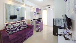 A modern living room with a purple sofa, a TV, and a kitchen area with a purple cabinet. - Comfort 2Br Apartment At Bassura City (Jakarta)