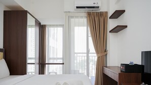 A hotel room with a bed, a desk, a television, an air conditioning unit, and a window with curtains. - Comfort And Warm Studio Room Apartment At M-Town Residence (Gading Serpong)
