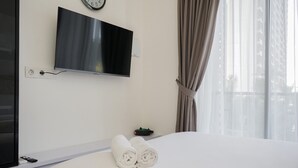 A hotel room with a bed, a TV mounted on the wall, a clock, and a window with curtains. - Elegant And Comfy Studio At Sky House Bsd Apartment (Pagedangan)