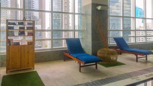 A modern indoor space with large windows, a wooden bookshelf, and two blue lounge chairs. - New Furnished And Comfortable 2Br At The Mayflower Apartment (Jakarta)