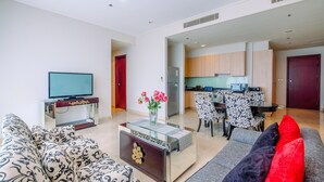 A modern living room with a sofa, a coffee table, and a TV. There is a kitchen area with a dining table and chairs visible through an open doorway. - New Furnished And Comfortable 2Br At The Mayflower Apartment (Jakarta)