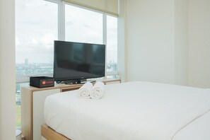 A hotel room with a bed, a flat-screen TV on a wooden stand, and a view of the city through large windows. - Minimalist And Comfort 1Br At B Residence (Pagedangan)