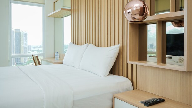A hotel room with a large bed, a wooden headboard, a television, and a city view. - Minimalist And Comfort 1Br At B Residence (Pagedangan)