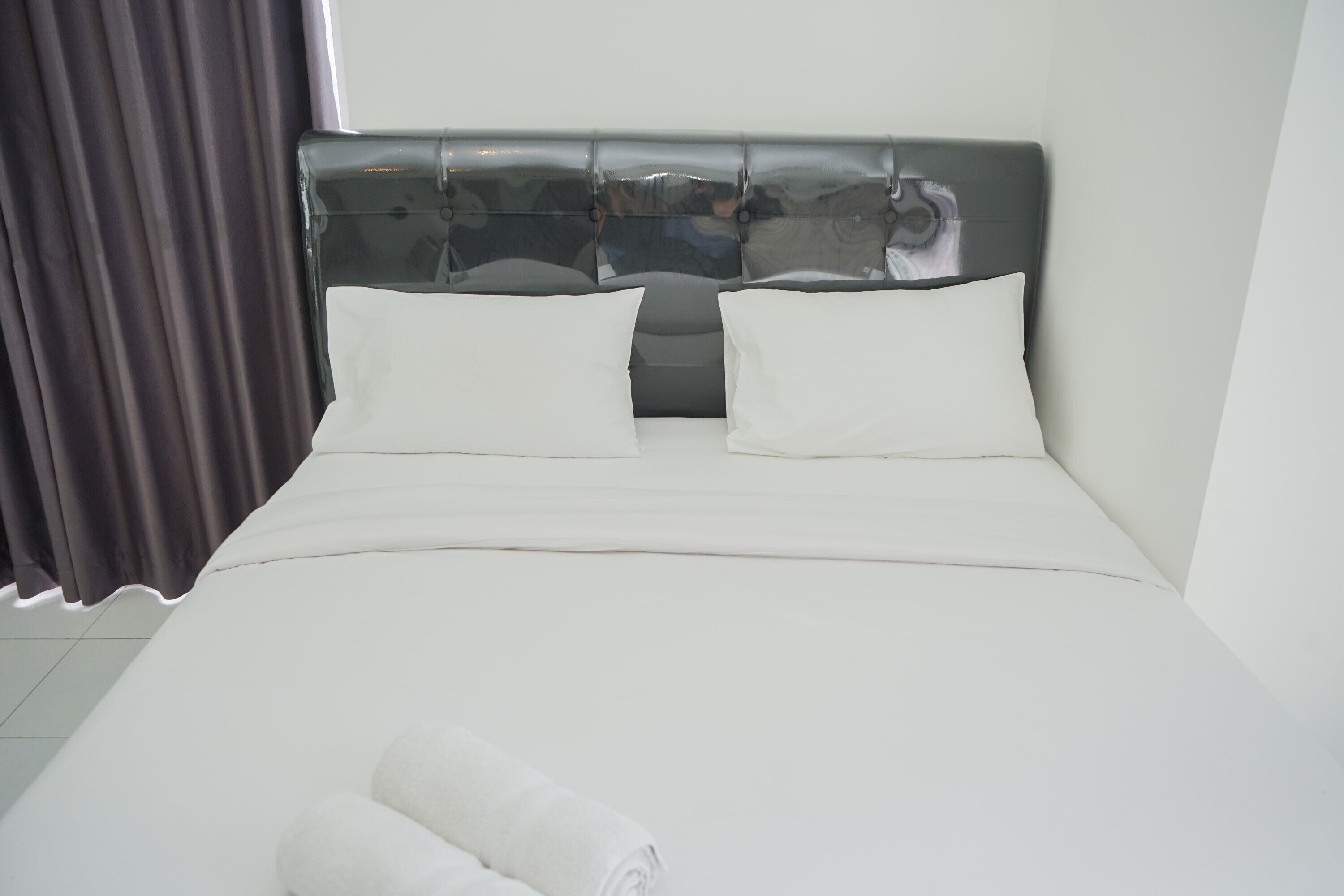 A neatly made bed with white linens and pillows, a grey upholstered headboard, and a dark curtain to the left.