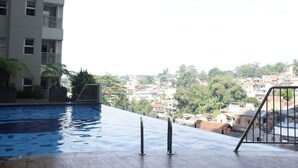An infinity pool with a wooden deck, surrounded by a high-rise building with balconies and a view of a densely populated area with trees.