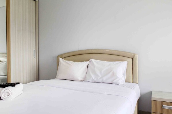 A neatly made bed with white bedding and pillows, a remote control, and a rolled towel on the left side.