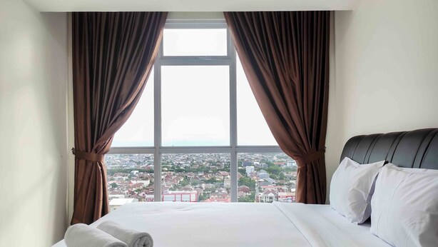 A hotel room with a large bed, white linens, and a view of a cityscape through a window with brown curtains. - Big And Comfy 3Br At Vida View Apartement (Makassar)