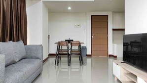 A modern living room with a grey sofa, a wooden dining table with chairs, a TV on a wooden stand, and a door with a light switch. - Big And Comfy 3Br At Vida View Apartement (Makassar)