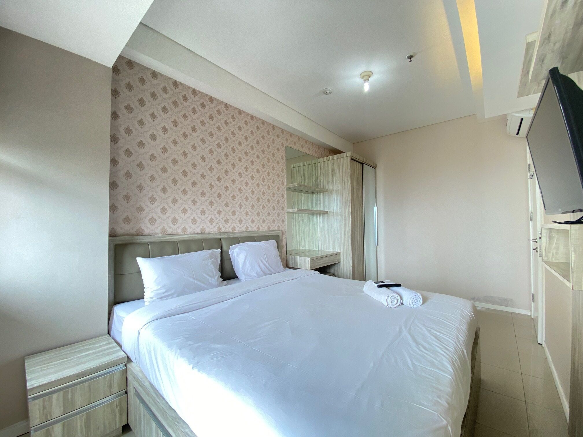 A neatly made bed with white linens, a wooden nightstand, a mirror, and a television on a stand.
