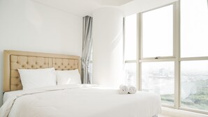 A large bed with a tufted headboard, white bedding, and a view of a cityscape through large windows. - Stunning And Strategic 2Br Gold Coast Apartment Near Pik (Jakarta)