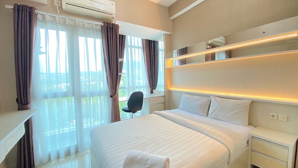 A hotel room with a large bed, a desk, and a chair. There is a window with curtains, an air conditioning unit, and a shelf with lights. - Cozy And Spacious Studio Apartment At Taman Melati Jatinangor (Hegarmanah)