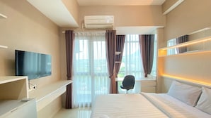 A hotel room with a bed, a desk, a chair, a TV, and an air conditioning unit. - Cozy And Spacious Studio Apartment At Taman Melati Jatinangor (Hegarmanah)