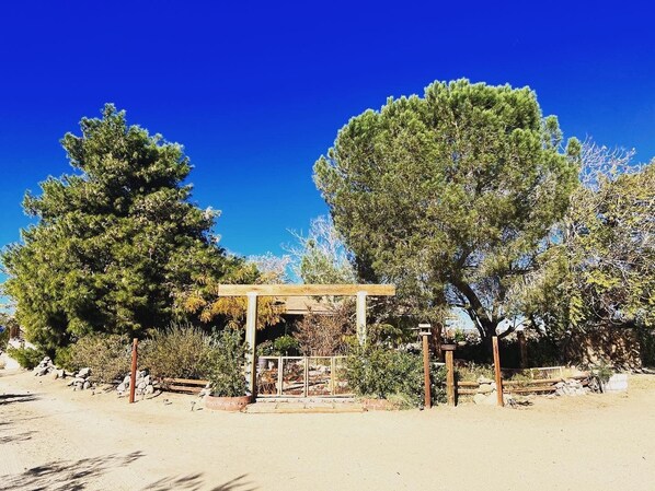A desert landscape with a wooden pergola structure, surrounded by large trees and sparse vegetation. - Ultimate relaxation on 4+ acres, hot tub, projector/game room, BBQ, stars, moon (Yucca Valley)