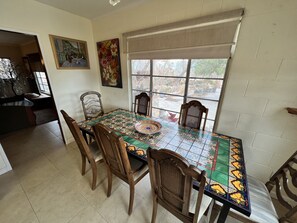 A dining area with a tiled table, wooden chairs, and a window with a view of the outdoors.