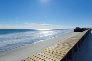 En træpromenade langs en strand med klar himmel og hav i baggrunden.