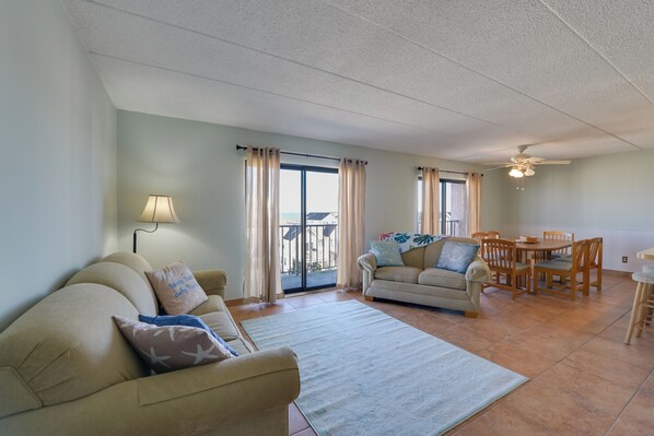 A living room with a sofa, a dining table with chairs, a ceiling fan, and a view through a glass door. - Simple North Wildwood Condo - Steps to Beach! (North Wildwood)