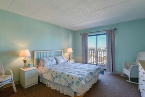 A bedroom with a bed, nightstands, a chair, a rocking chair, and a view through a window. - Simple North Wildwood Condo - Steps to Beach! (North Wildwood)