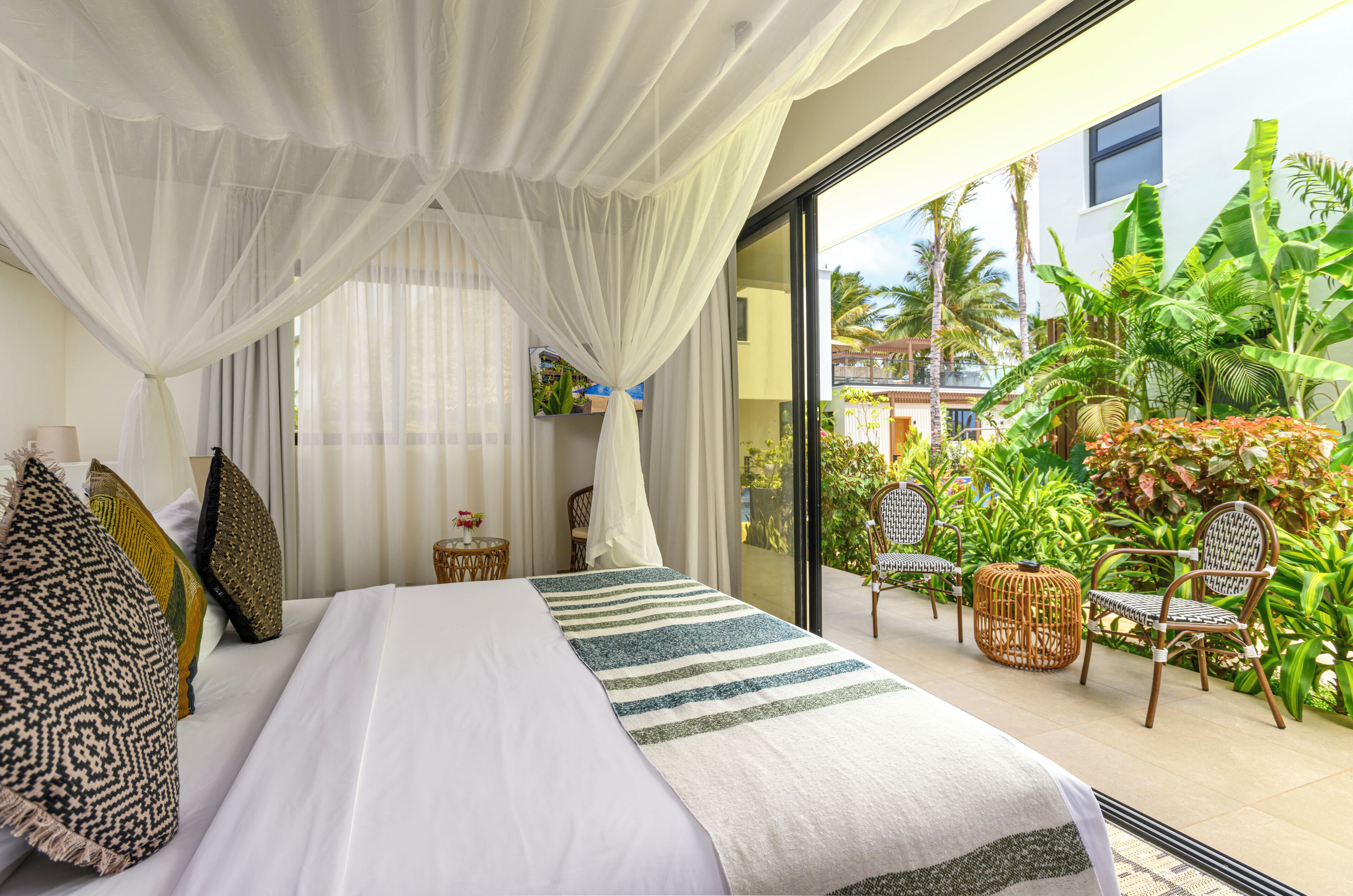 A bedroom with a large bed, white curtains, and a view of a tropical garden through a glass door.