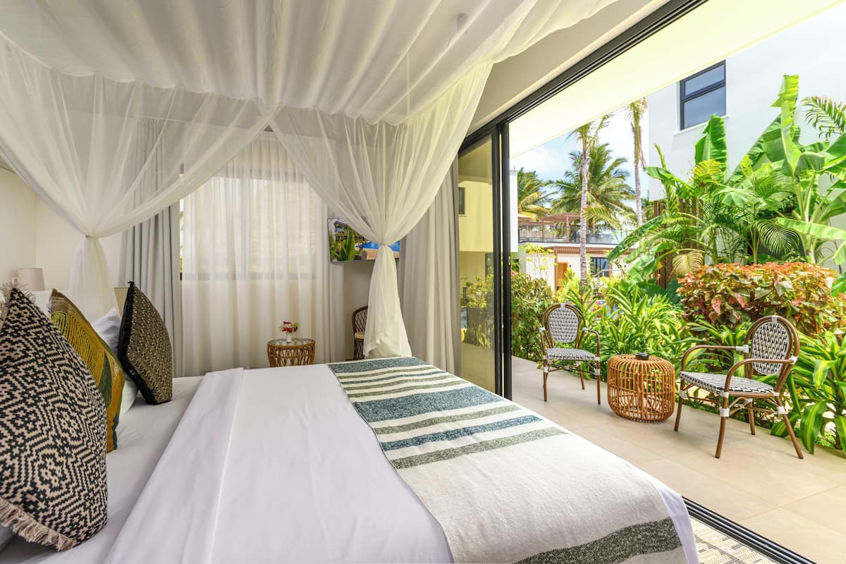 A bedroom with a large bed, white curtains, and a view of a tropical garden through a glass door.